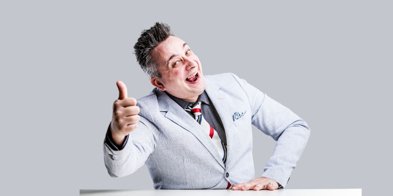 A white man with greying hair light grey suit gives a thumbs up to the camera while posing in a comedic manner